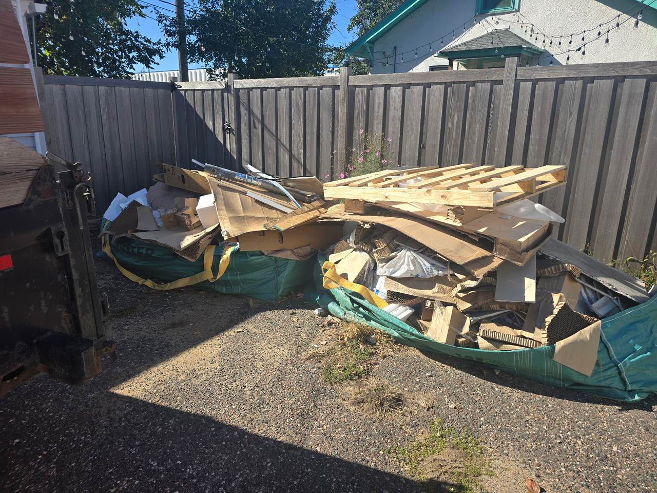 Two loaded Bagsters of cardboard, pallets and lumber against a backyard fence, beside a dump trailer