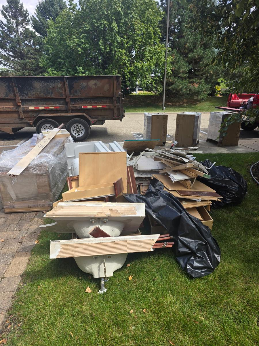 Torn-out cabinets, two sinks and broken drywall stacked on a lawn, with a loaded dump trailer behind