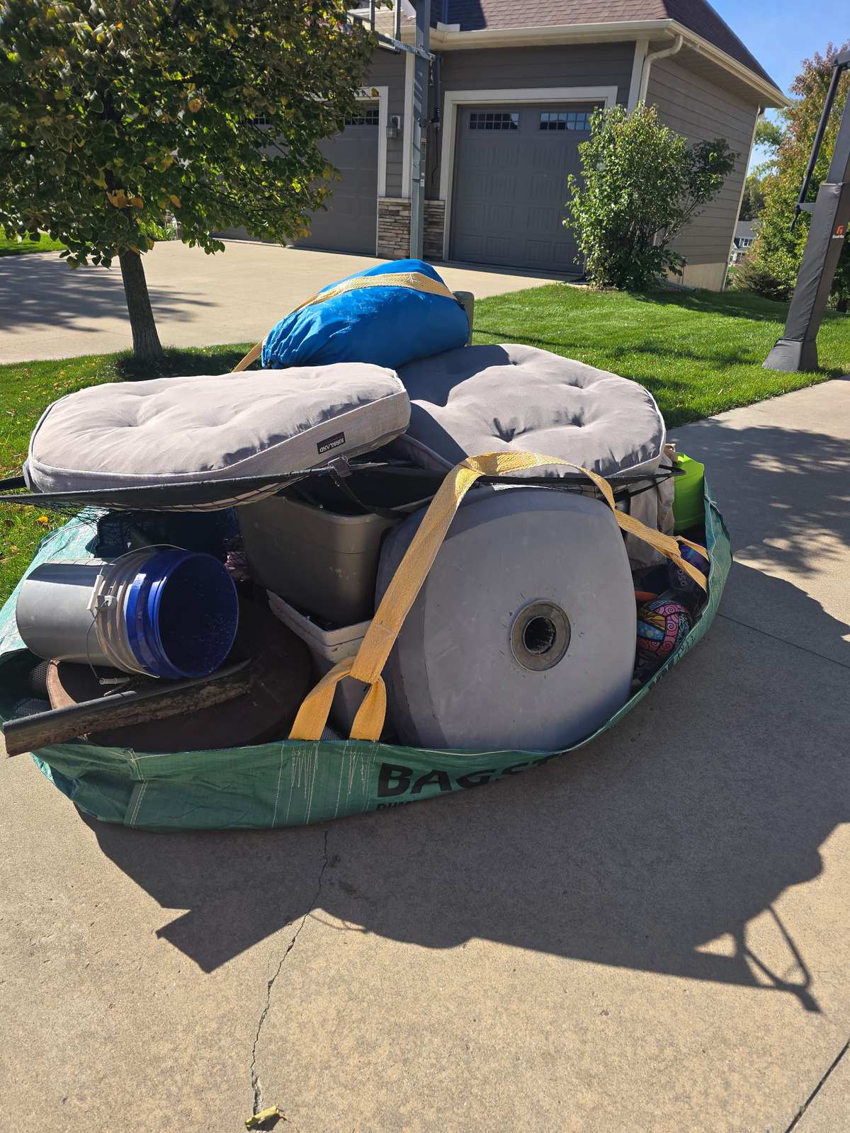A Bagster on a driveway loaded with patio cushions, storage bins, a bucket and household junk