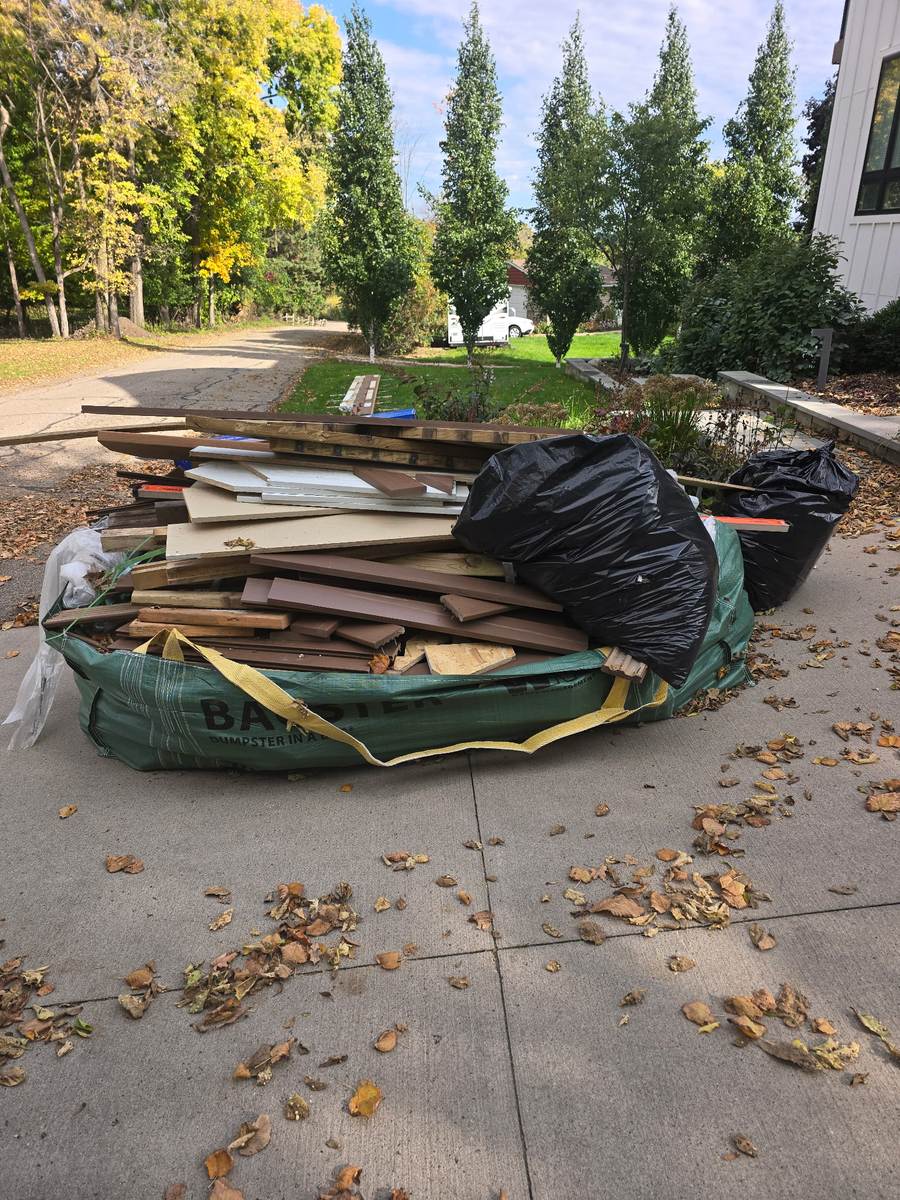 A Bagster on a driveway piled high with deck boards, lumber offcuts and two trash bags