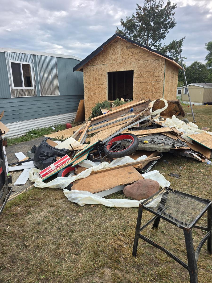 A torn-down shed piled on the grass beside a mobile home: roofing, framing, plywood and plastic sheeting